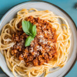 Een bord met spaghetti bolognese gegarneerd met een blaadje basilicum, naast twee eiwitrepen en verse prei op een blauwe houten tafel.